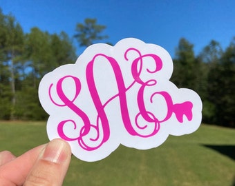 May include: A white sticker with a pink monogram "SAE" and a pink tooth graphic. The sticker is held in a hand against a blurred background of green grass and a blue sky.