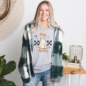 May include: A light gray t-shirt with a ghost graphic and the words "Stay Spooky" in orange. The shirt is worn with a green and white plaid flannel shirt and blue jeans. The ghost graphic is centered on the shirt.