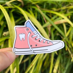 May include: Pink high-top sneaker with a white lace and a blue tongue. The sneaker has a white outline and a white tooth graphic on the side.