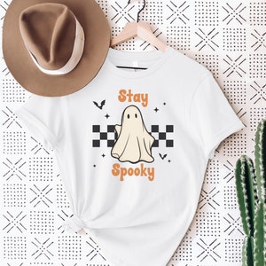 May include: White t-shirt with a Halloween-themed design. The shirt features a cartoon ghost with the words "Stay Spooky" in orange. The ghost is set against a black and white checkered background, with bats and stars.
