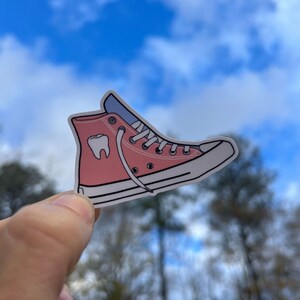May include: Pink high-top sneaker with a white sole and a white tooth graphic on the side. The sneaker is held in a hand against a blue sky background.