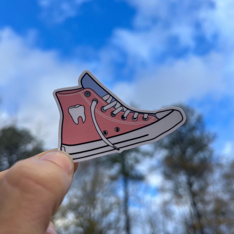 May include: Pink high-top sneaker with a white sole and a white tooth graphic on the side. The sneaker is held in a hand against a blue sky background.