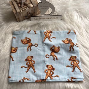 May include: A light blue fabric featuring a repeating pattern of cartoon monkeys. A yellow measuring tape is next to the fabric. The image also includes a wooden box with seashells and a decorative sign that reads "Coole Kleidung-coole Kids" and "StoffoaseKreationen."