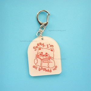 May include: A white acrylic keychain with a red outline of a cat wearing glasses and reading a book. The text "Sorry, I'm all booked up" is printed in red on the keychain.