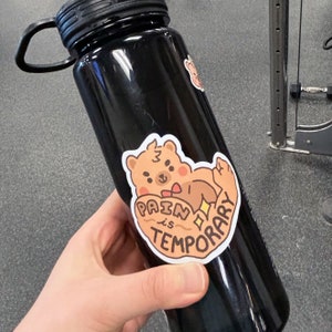 Puede incluir: Botella de agua negra con una pegatina blanca que muestra un oso de dibujos animados con una pajarita roja y el texto "Pain is Temporary".