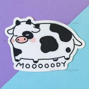 May include: A white and black cartoon cow with a pink nose and the word "MOOOOODY" written below it.