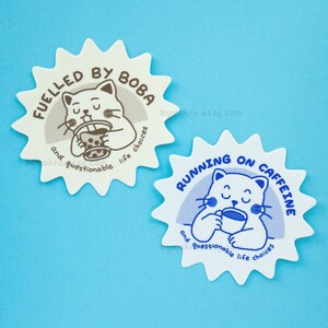 May include: Two white star-shaped stickers with blue and brown outlines. The sticker on the left features a cartoon cat drinking boba tea with the text "FUELLED BY BOBA and questionable life choices". The sticker on the right features a cartoon cat drinking coffee with the text "RUNNING ON CAFFEINE and questionable life choices".