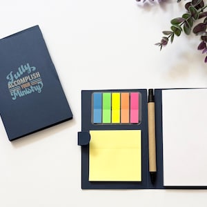 May include: A dark blue folding stationery set featuring a closed notebook with the text "Fully ACCOMPLISH YOUR Ministry" and an open notebook containing a yellow square sticky note, five colorful page marker tabs, and a brown pen.