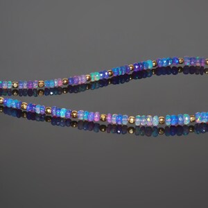 May include: A delicate necklace featuring small, faceted opal beads in shades of blue, purple, and pink. Gold-coloured beads are interspersed throughout the necklace. The necklace has a gold-coloured clasp and chain.