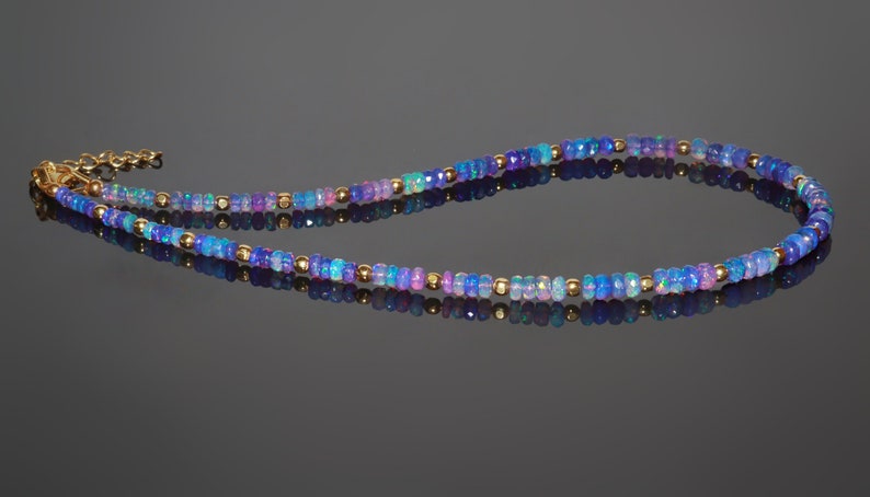 May include: A delicate necklace featuring small, faceted opal beads in shades of blue, purple, and pink. Gold-coloured beads are interspersed throughout the necklace. The necklace has a gold-coloured clasp and chain.