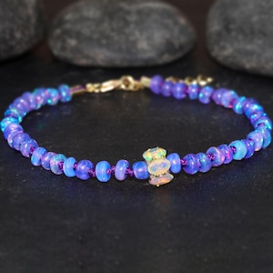 May include: A delicate bracelet made with small, iridescent purple opal beads and a larger, white opal bead in the center. The bracelet is strung on a purple cord and has a gold clasp.