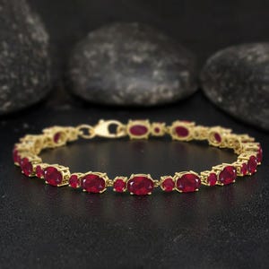 May include: A gold bracelet featuring oval-cut, deep red gemstones. The bracelet is set in a gold chain with a secure clasp. The gemstones are evenly spaced along the length of the bracelet, creating a luxurious look.