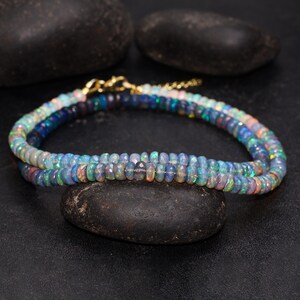 May include: A double-strand necklace composed of small, iridescent opal beads. The beads display a range of colors, including blues, greens, and hints of pink and orange. The necklace is secured with a gold-colored clasp and chain.