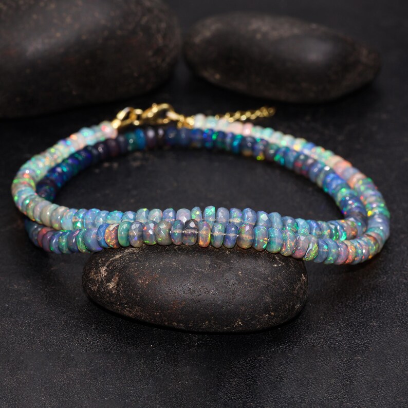 May include: A double-strand necklace composed of small, iridescent opal beads. The beads display a range of colors, including blues, greens, and hints of pink and orange. The necklace is secured with a gold-colored clasp and chain.