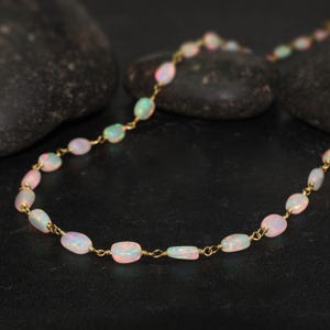 Op de afbeelding: Delicate ketting met ovale opalen aan een gouden ketting. De opalen vertonen een zachte roze tint en iriserende kleuren. De ketting wordt gepresenteerd tegen een donkere achtergrond, wat het fijne ontwerp en de kleuren benadrukt.