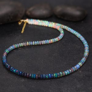 May include: A multi-colored opal bead necklace with a gold-colored chain clasp. The beads transition from dark blue to lighter shades of blue, green, and red. The necklace is displayed on a black surface.