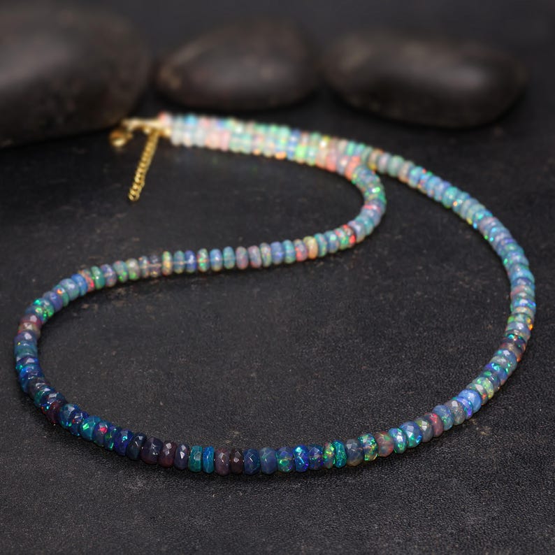 May include: A multi-colored opal bead necklace with a gold-colored chain clasp. The beads transition from dark blue to lighter shades of blue, green, and red. The necklace is displayed on a black surface.