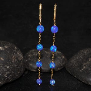 May include: A pair of gold-colored dangle earrings. Each earring features a series of blue opal beads connected by a delicate gold chain. The earrings have a lever-back closure. The opal beads have a shimmering, iridescent quality.