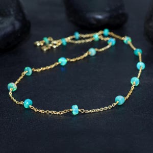 May include: A delicate gold chain necklace with small, round, light blue opal beads. The necklace is on a dark background.