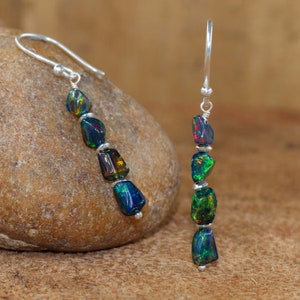May include: Dangle earrings with black opal stones, showing flashes of green, blue, and red. The opals are strung on silver-coloured wire. The earrings are displayed on a brown stone.