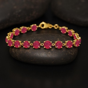 Peut inclure: Un bracelet en or orné de pierres précieuses rouges vives de forme ovale. Les pierres sont serties dans des montures en or individuelles et reliées entre elles. Le bracelet est présenté sur une surface sombre et texturée.