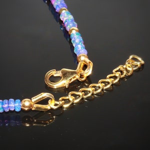 May include: Close-up of a necklace featuring blue and purple opal beads and gold detailing. The necklace has a gold clasp and an extender chain. The beads are small and faceted, with shiny gold elements.