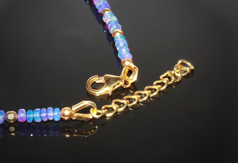 May include: Close-up of a necklace featuring blue and purple opal beads and gold detailing. The necklace has a gold clasp and an extender chain. The beads are small and faceted, with shiny gold elements.