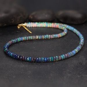 May include: A beaded necklace composed of small, iridescent opal stones. The opals display a range of colors, including blues, greens, and pinks, with a gold-colored clasp. The necklace is displayed on a dark surface.