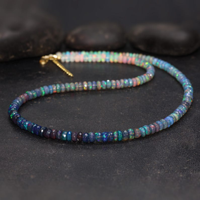 May include: A beaded necklace composed of small, iridescent opal stones. The opals display a range of colors, including blues, greens, and pinks, with a gold-colored clasp. The necklace is displayed on a dark surface.