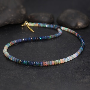 May include: A beaded necklace composed of small, colorful opal stones. The necklace features a gold-colored clasp and is displayed on a dark surface with black stones in the background. The opal beads display a range of colors.