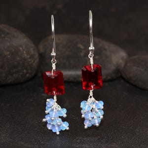 May include: A pair of dangle earrings featuring a rectangular, deep red gemstone at the top, connected to a cluster of small, light blue opalescent beads. The earrings have silver-colored hooks.