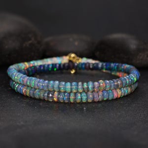 May include: Two beaded bracelets with iridescent opal beads. The beads are a mix of blues, greens, and pinks, strung together with a gold clasp. The bracelets are displayed on a dark surface with black stones in the background.