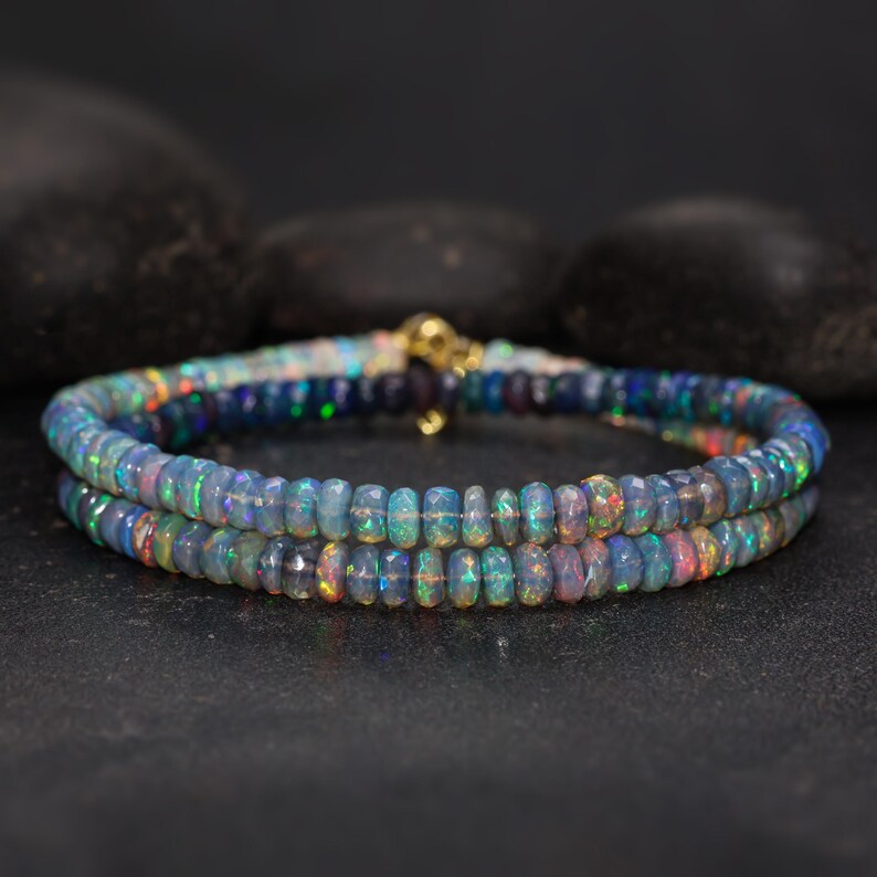 May include: Two beaded bracelets with iridescent opal beads. The beads are a mix of blues, greens, and pinks, strung together with a gold clasp. The bracelets are displayed on a dark surface with black stones in the background.
