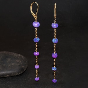 Könnte beinhalten: Ein Paar lange, goldene Ohrringe mit violetten Opalperlen. Die Ohrringe haben eine zarte Kette und sind perfekt, um jedem Outfit einen Hauch von Eleganz zu verleihen.