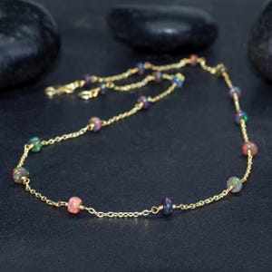 Peut inclure: Un collier en chaîne dorée avec de petites perles rondes d'opale multicolores. Le collier est sur une surface gris foncé.