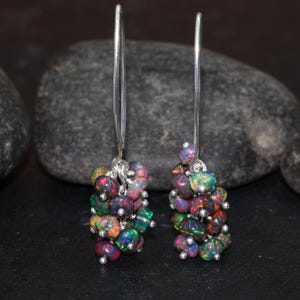 May include: A pair of silver dangle earrings with a cluster of iridescent black opal stones. The earrings are made with a simple silver hoop and a long silver chain that hangs down to the cluster of stones.