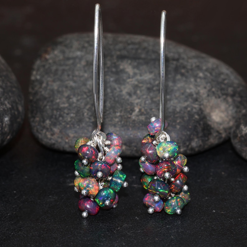 May include: A pair of silver dangle earrings with a cluster of iridescent black opal stones. The earrings are made with a simple silver hoop and a long silver chain that hangs down to the cluster of stones.