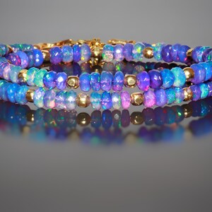 May include: Three-strand bracelet with iridescent blue opal beads and gold-coloured accents. The opal beads display a range of colours, including blue, purple, and green. The bracelet is designed with small, round gold-coloured beads between the opal beads.