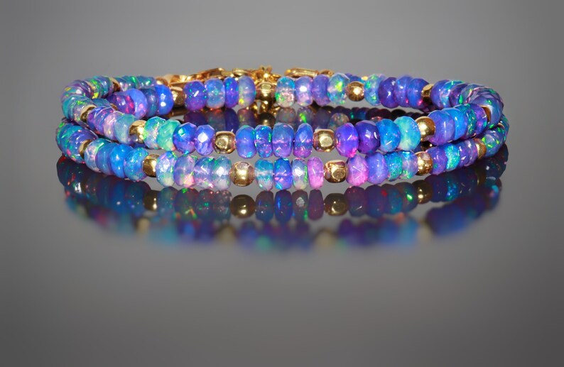 May include: Three-strand bracelet with iridescent blue opal beads and gold-coloured accents. The opal beads display a range of colours, including blue, purple, and green. The bracelet is designed with small, round gold-coloured beads between the opal beads.