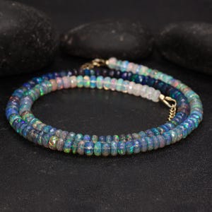 May include: A layered necklace featuring iridescent opal beads in shades of blue, green, and white. The beads are faceted and strung together with gold-colored clasps. The necklace is displayed on a black surface.
