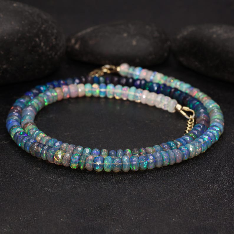 May include: A layered necklace featuring iridescent opal beads in shades of blue, green, and white. The beads are faceted and strung together with gold-colored clasps. The necklace is displayed on a black surface.
