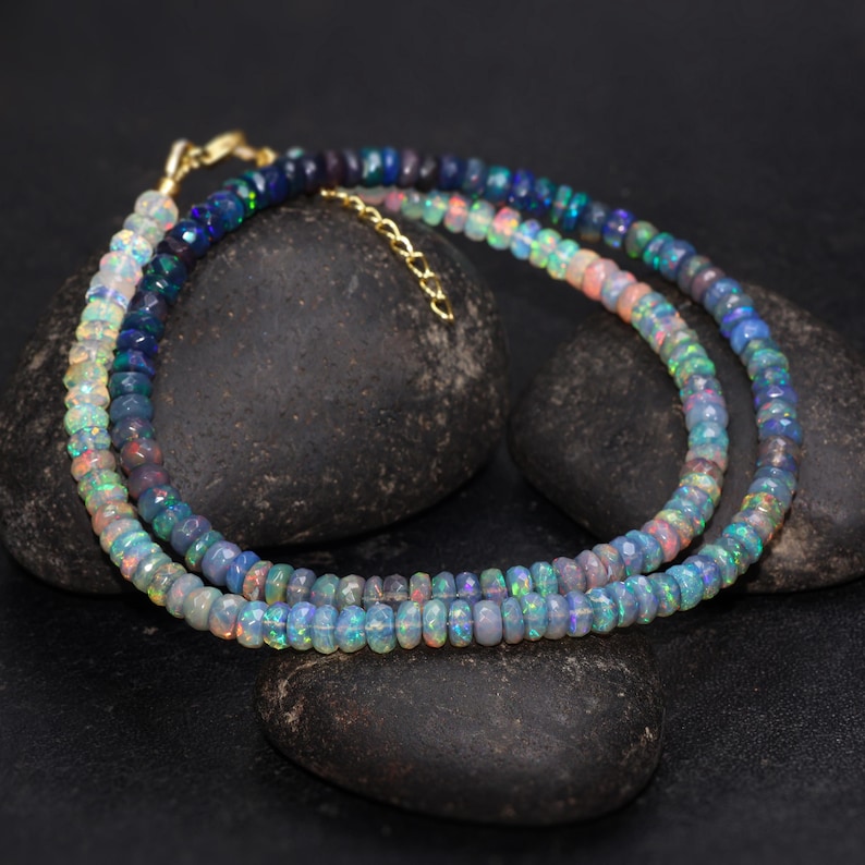 May include: Two necklaces made of small, iridescent opal beads. The beads display a range of colors, including blues, greens, and pinks. The necklaces are draped over dark, textured stones, with a gold-colored clasp.