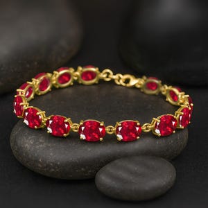 May include: A gold bracelet with oval, bright red gemstones. Each gemstone is set in a gold bezel and linked by a fine gold chain. The bracelet is presented against a dark backdrop, highlighting the jewellery's colour.