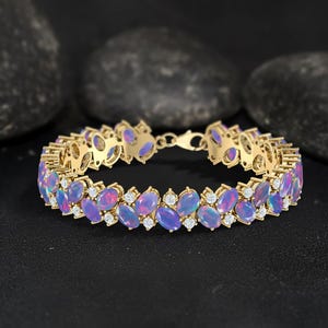 May include: A gold bracelet featuring oval-shaped opals in shades of purple and pink, interspersed with small, round diamonds. The bracelet is set against a dark background, highlighting the gemstones' colors.