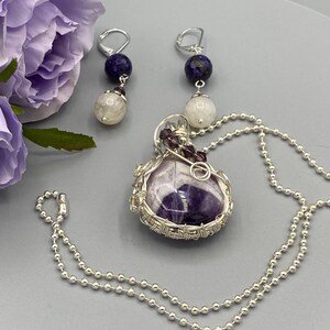 May include: A silver chain necklace with a purple amethyst stone pendant wrapped in silver wire. The pendant is accented with small purple beads. The necklace comes with a pair of earrings with a silver hook, a purple stone, and a white stone.