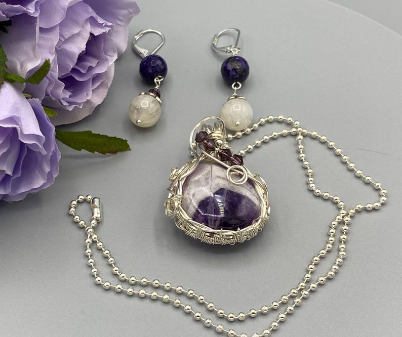 May include: A silver chain necklace with a purple amethyst stone pendant wrapped in silver wire. The pendant is accented with small purple beads. The necklace comes with a pair of earrings with a silver hook, a purple stone, and a white stone.