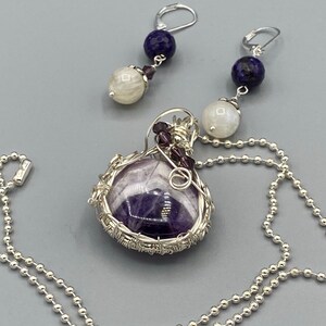 May include: A silver chain necklace with a purple amethyst stone pendant wrapped in silver wire. The pendant also features small purple and white beads. The necklace comes with a pair of matching earrings with a white moonstone and a blue lapis lazuli bead.