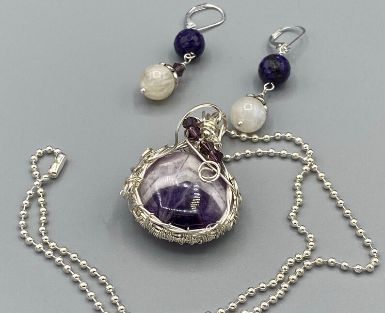 May include: A silver chain necklace with a purple amethyst stone pendant wrapped in silver wire. The pendant also features small purple and white beads. The necklace comes with a pair of matching earrings with a white moonstone and a blue lapis lazuli bead.