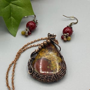 May include: A copper wire wrapped pendant necklace with a red, yellow, and brown agate stone. The necklace comes with a pair of matching earrings with red agate stones.