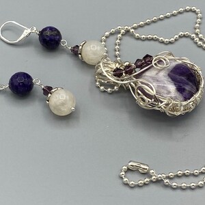 May include: A silver necklace with a purple and white amethyst stone pendant wrapped in silver wire. The necklace has a silver chain and a silver clasp. The pendant is surrounded by small purple beads. The necklace comes with a pair of earrings with a blue stone, a white stone, and a purple bead.